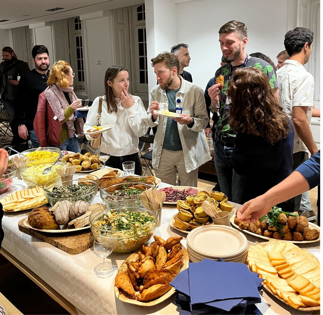 A number of people talking and eating in front of a table brimming with a delicious assortment of food.